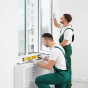 Two men in green overalls work on installing a window in a white room.