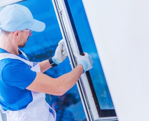 A man in a blue outfit and overalls installs a replacement window in a white room.
