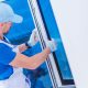 A man in a blue outfit and overalls installs a replacement window in a white room.