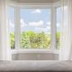 A closeup image of a bay window in a bedroom. You can see the gray wallpaper, white curtains, and the trees and skyline outside the house.