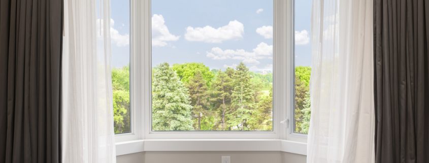 A closeup image of a bay window in a bedroom. You can see the gray wallpaper, white curtains, and the trees and skyline outside the house.