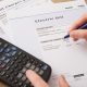 A closeup of a calculator next to a stack of papers on a desk. A person's hand holding a pen hovers above a sheet of paper that reads "electric bill".