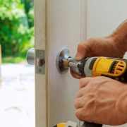 A worker's hands are shown operating a yellow drill on an entry door. They are drilling the knob into place here.