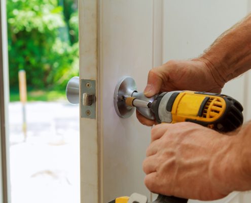 A worker's hands are shown operating a yellow drill on an entry door. They are drilling the knob into place here.