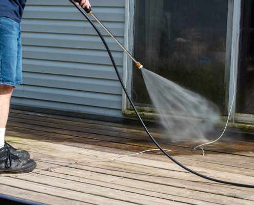 A man in jean shorts uses a power washer to wash grime and mold off of a patio door. You can see the wooden deck he is standing on and the blue finish of the home's exterior.