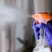 A purple-gloved hand squeezes down on a clear squirt bottle with an orange top to clean the clear glass in front of it. You can see the cleaning liquid shooting from the end of the spray bottle.