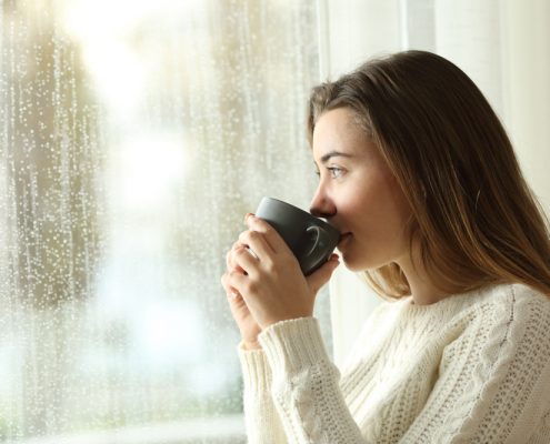 A woman sips out of a brown coffee mug while looking out of a window. You can see a plethora of raindrops running down the pane.