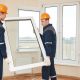 Two men in orange hard hats and blue suits move a window pane into position to be installed on the beige wall behind them.