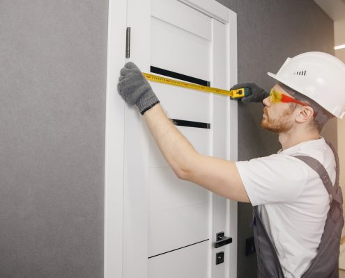 A carpenter in a white hard hat and overalls measures the width of a white door in a grey room.