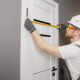 A carpenter in a white hard hat and overalls measures the width of a white door in a grey room.